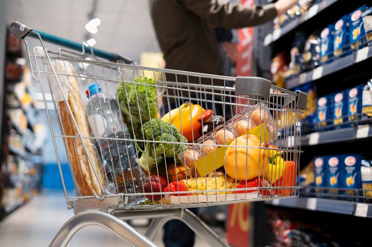 Una persona sceglie dei prodotti da uno scaffale del supermercato dietro un carrello della spesa pieno