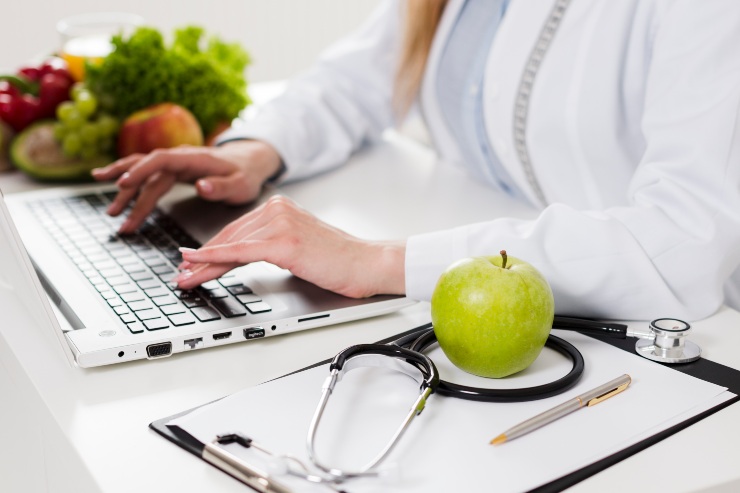 Un medico scrive al pc seduto alla scrivania, accanto a una mela, uno stetoscopio e un blocco