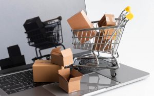 Laptop aperto su una scrivania con un modellino di carrello della spesa e pacchi di cartone