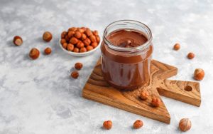Un barattolo di crema spalmabile alla nocciola su un tagliere di legno vicino a una ciotola di nocciole